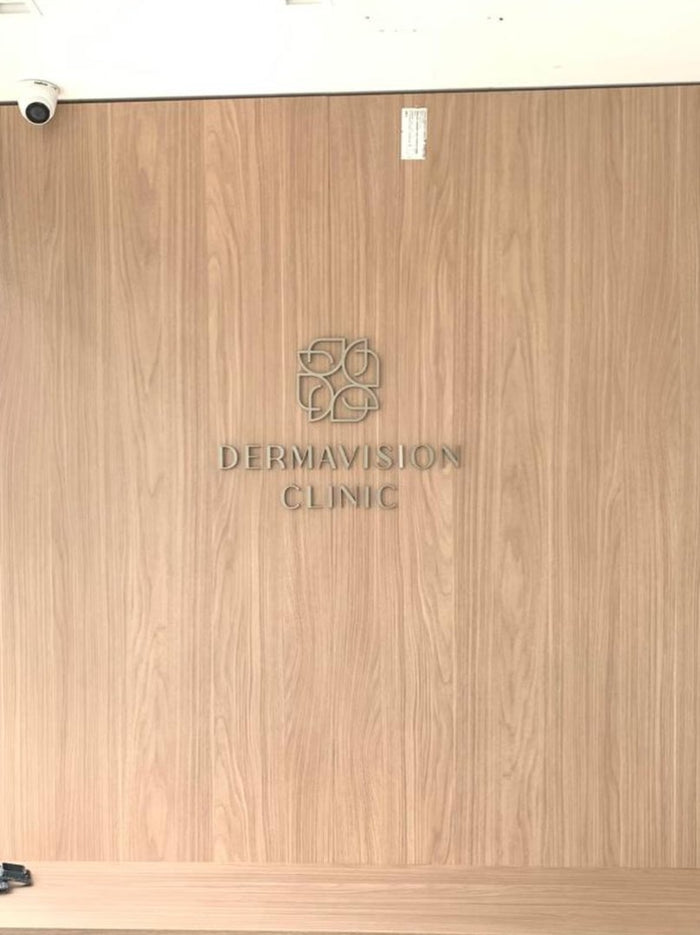 Dermavision Clinic: Logo em metal escovado sobre painel de madeira.