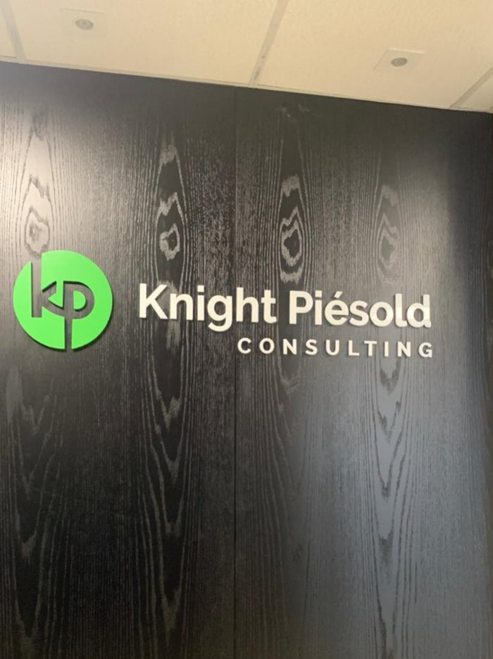 Knight Piésold Consulting: Logo em alto relevo com símbolo em verde, aplicada em painel de madeira escura.