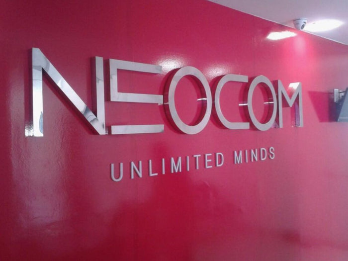 Neocom: Logomarca em letra caixa de aço inox polido sobre parede colorida.